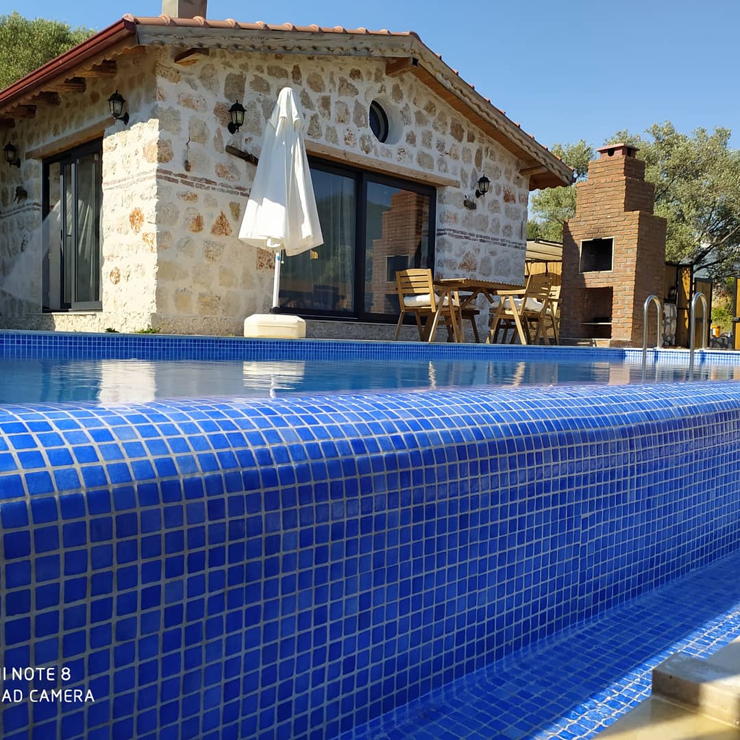 A clear view of a serene outdoor pool is presented, featuring blue tiles and gentle reflections of the sky. The pool is adjacent to a rustic stone house with a welcoming patio area. A white umbrella provides shade over a small dining set made of wood.
