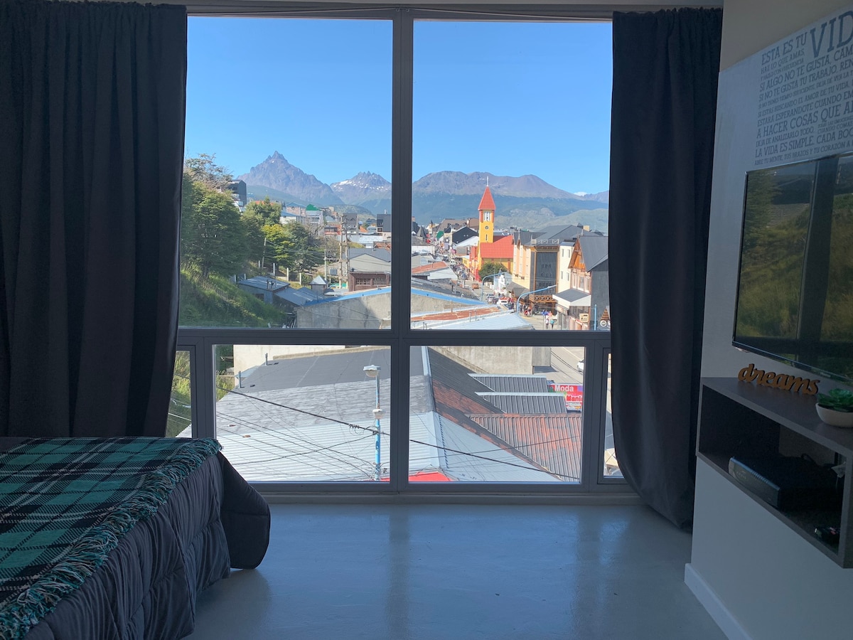 A spacious window provides an expansive view of Ushuaia's vibrant cityscape and surrounding mountains. The room features a cozy bed layered with a checked blanket, while dark curtains frame the window, enhancing the scenic outlook on a clear day.
