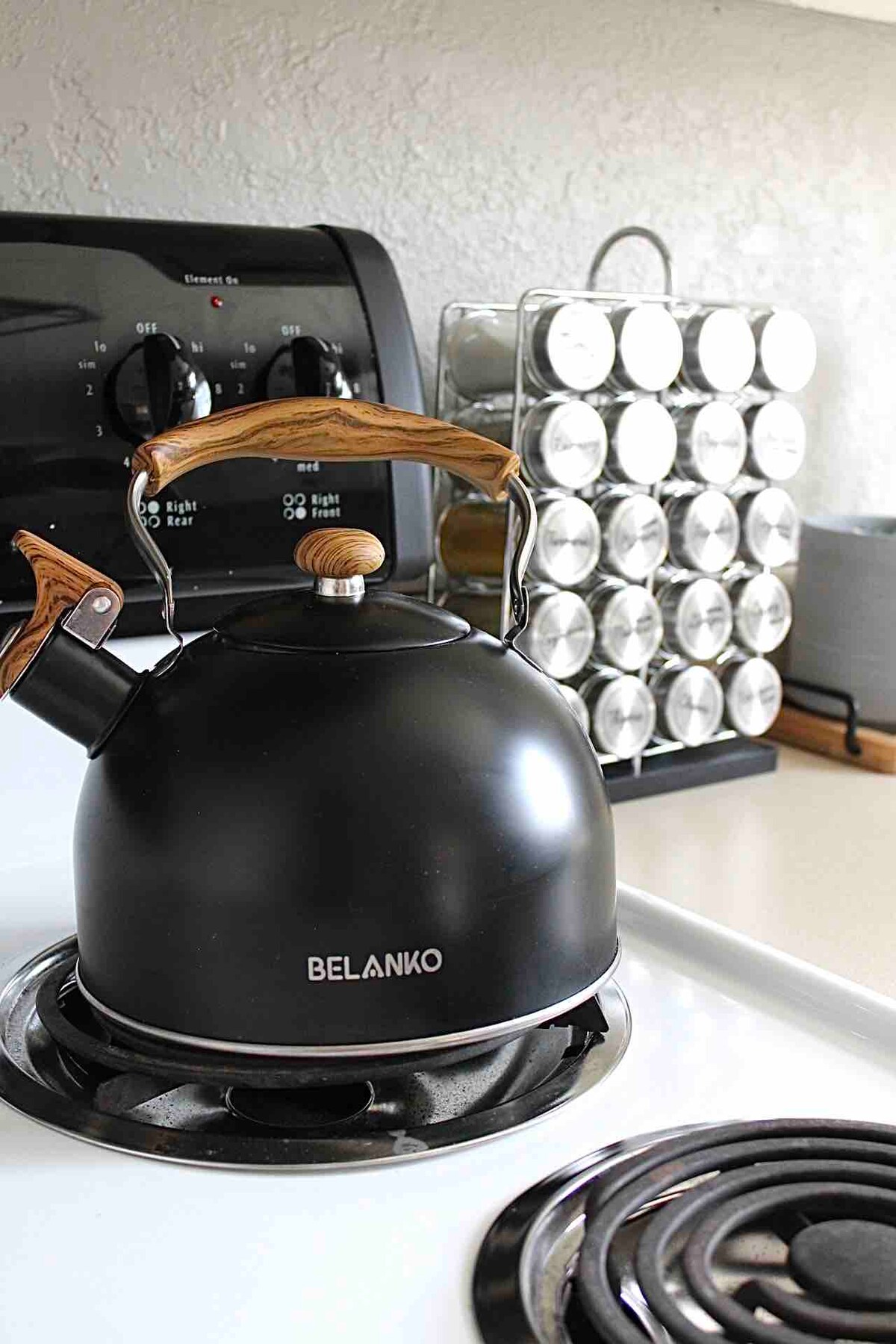 A black kettle with a wooden handle sits atop a white stove. Behind it, a rack of spice containers is organized neatly, contributing to a functional kitchen space.
