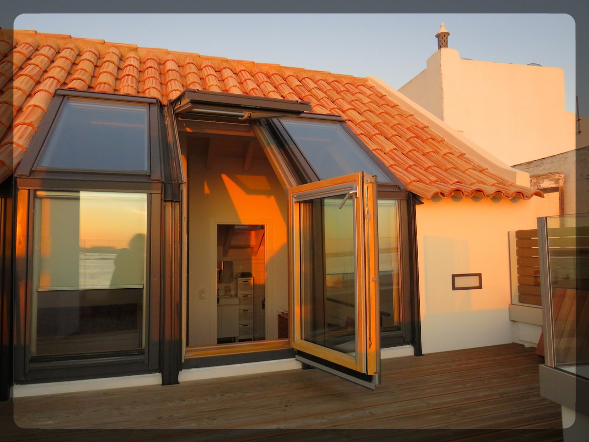 A modern rooftop terrace is featured, showcasing large glass doors that open outward, allowing for natural light to fill the space. The red-tiled roof contrasts with the warm glow of sunset, offering a glimpse of the serene surroundings.