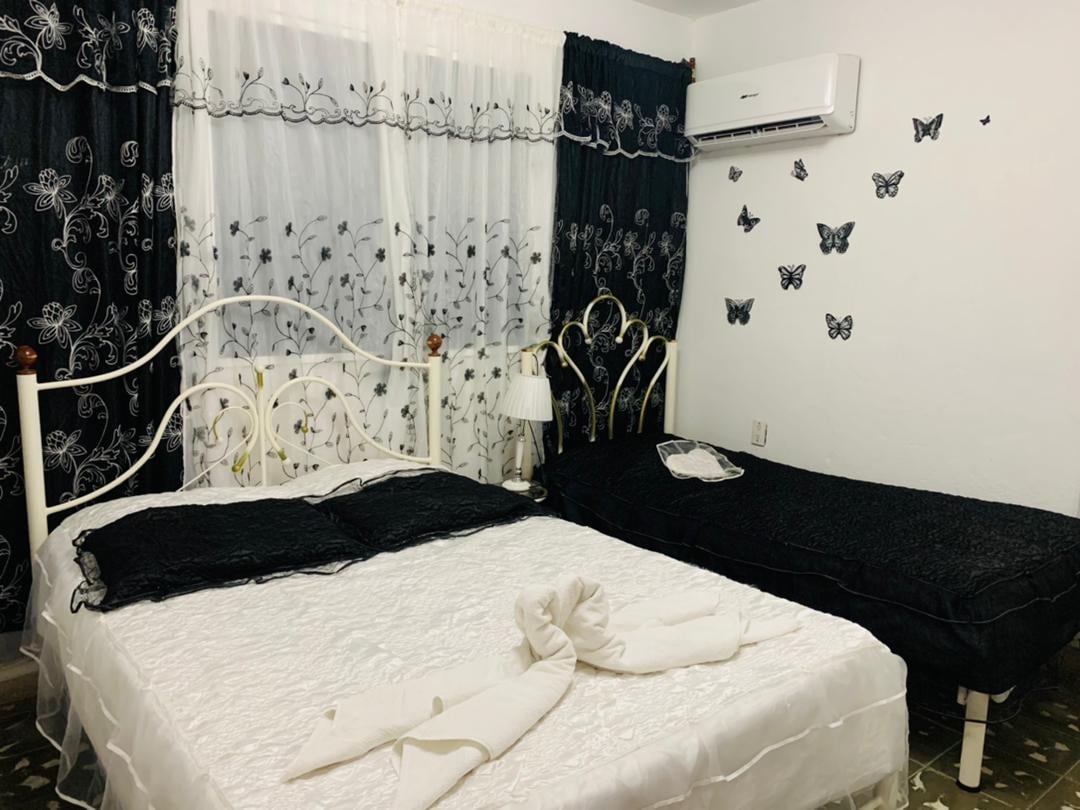 A bright bedroom is equipped with a spacious bed and an additional twin bed. Black and white decor features delicate butterfly wall art and sheer curtains, allowing soft light to fill the room. An air conditioning unit provides comfort in the space.