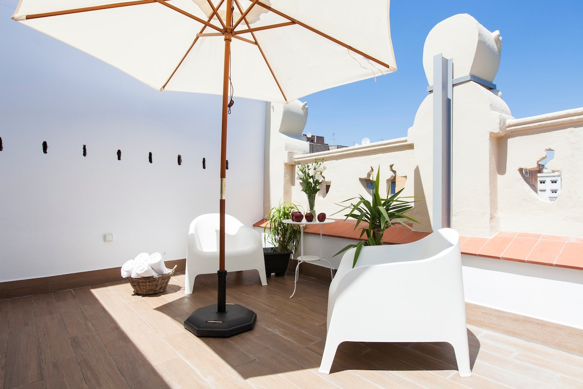 A private terrace is presented, featuring two modern white chairs under a large umbrella. Potted plants add greenery to the space, and a small basket with towels is neatly arranged, creating an inviting area for relaxation.