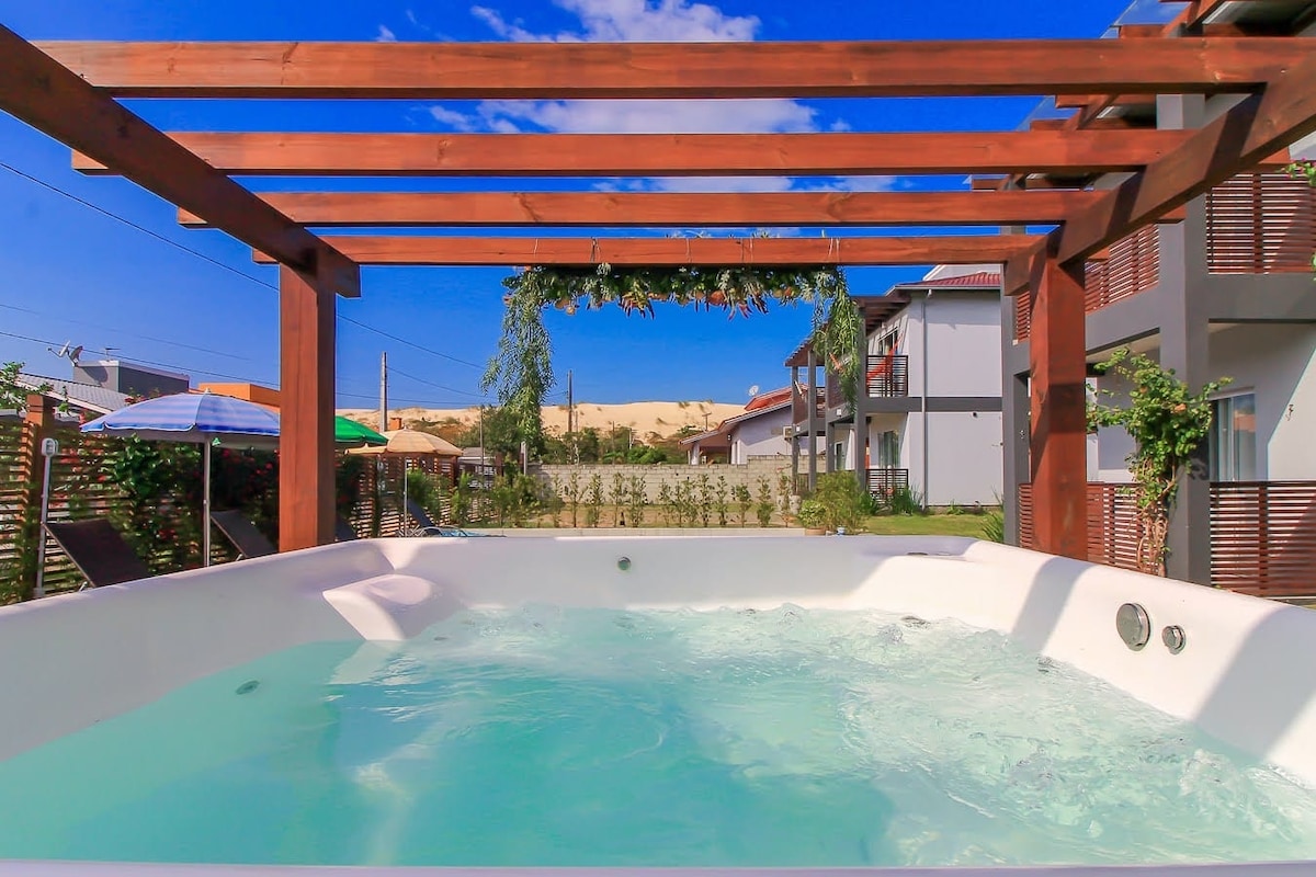 A calming outdoor spa is surrounded by a wooden pergola, featuring a clear, bubbly water surface. In the background, sun loungers and umbrellas provide shade, while lush greenery adds a natural touch to the tranquil space.