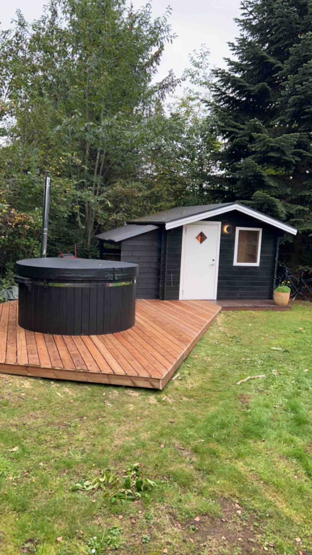 A wooden deck extends around a round black hot tub, which sits adjacent to a small cabin. The cabin features a white door and a diamond-shaped window, surrounded by lush greenery and trees, contributing to a private and serene outdoor setting.