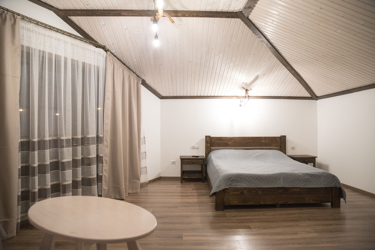 A spacious room features a large wooden bed centered beneath a vaulted ceiling, complemented by soft lighting. Light-colored curtains frame a window, while a simple round table rests on the polished wood floor, enhancing the room's open atmosphere.