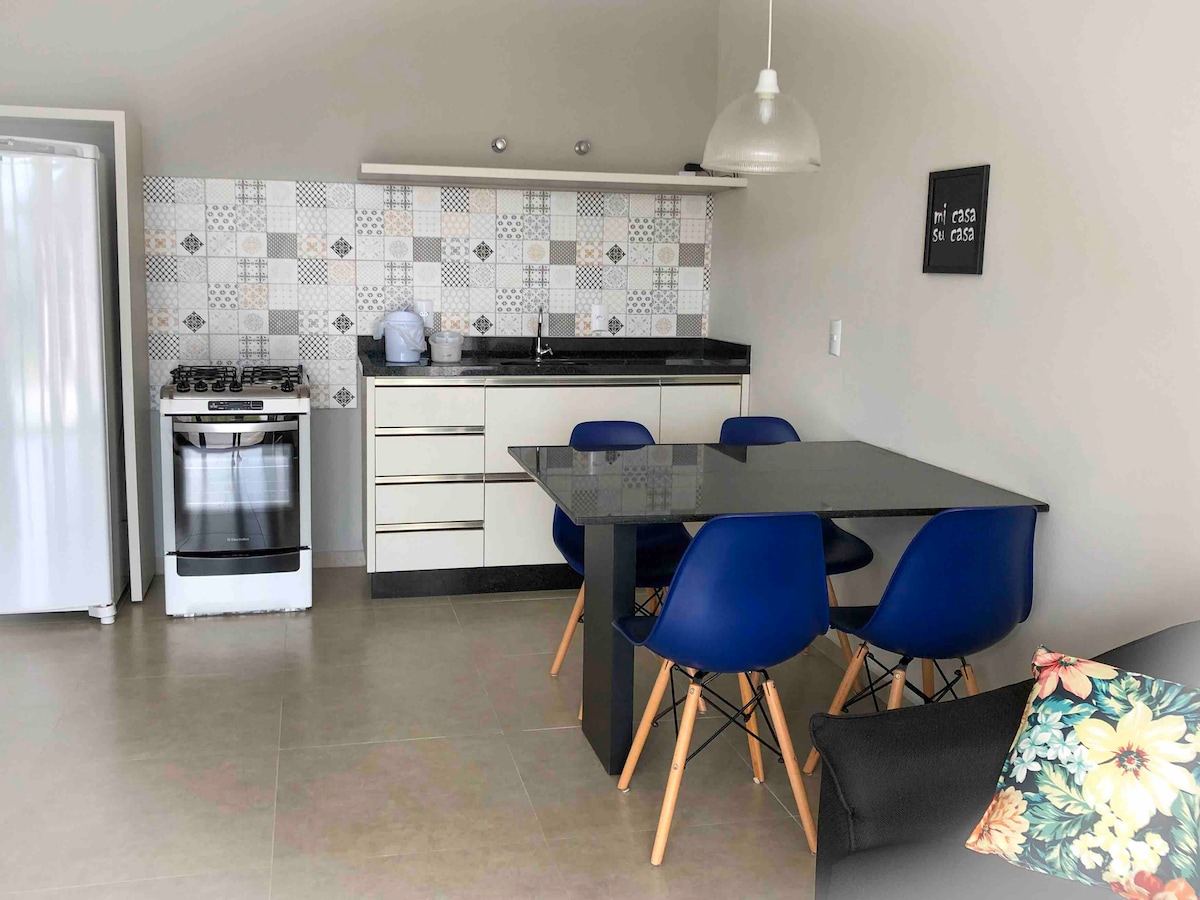 A modern kitchen area features a sleek black countertop and a four-seat dining table with blue chairs. Stainless steel appliances include a gas stove and refrigerator, set against a stylish patterned backsplash. Natural light fills the space, creating an inviting atmosphere.