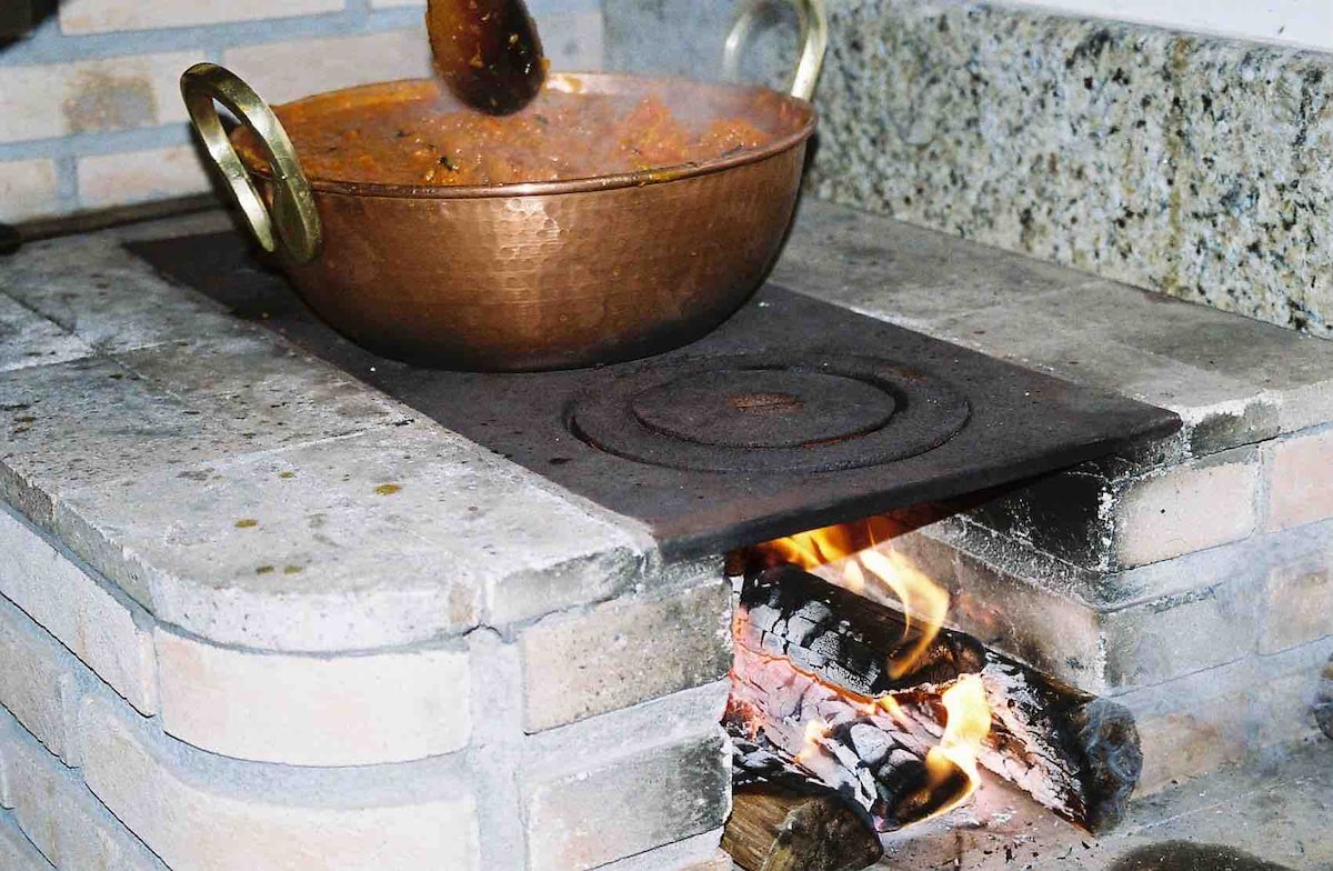 A copper pot filled with food is seen resting on a stone countertop, with a traditional fireplace visible underneath. Flames can be observed dancing around pieces of wood, providing warmth and a cooking element to the rustic kitchen setting.