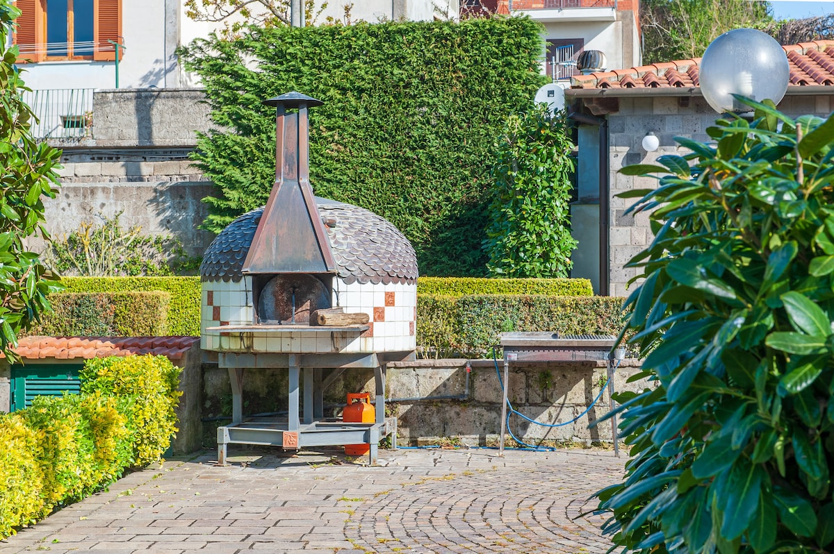 An outdoor area is presented, featuring a traditional stone pizza oven with a distinctive tiled exterior. Lush green hedges surround the space, while a barbecue grill is positioned nearby. Circular stone pavers create a welcoming and functional area for outdoor cooking and gatherings.