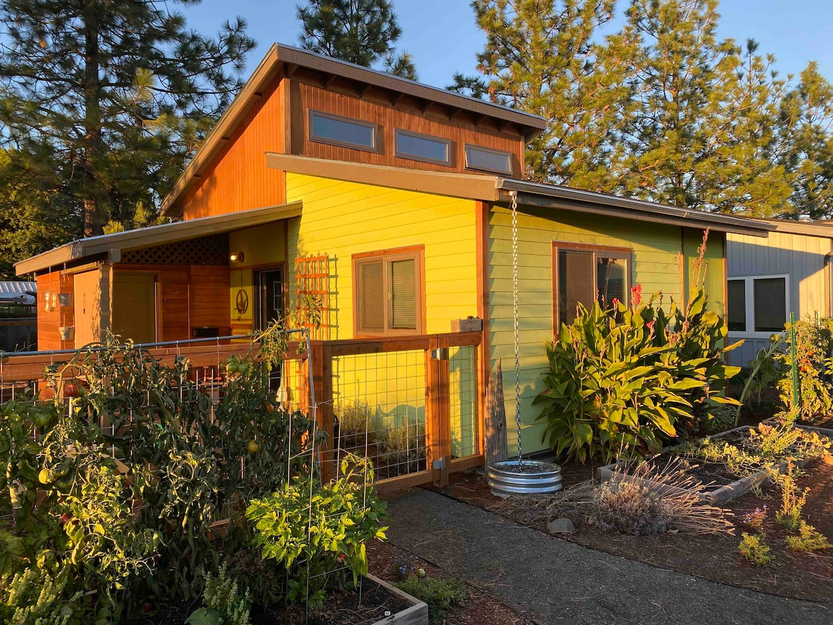 The exterior of a vibrant yellow house is framed by lush greenery and a vegetable garden, inviting guests to explore. Skylights and transom windows enhance the natural light, while a fenced area offers privacy. A pathway leads to the entrance, surrounded by flowering plants.