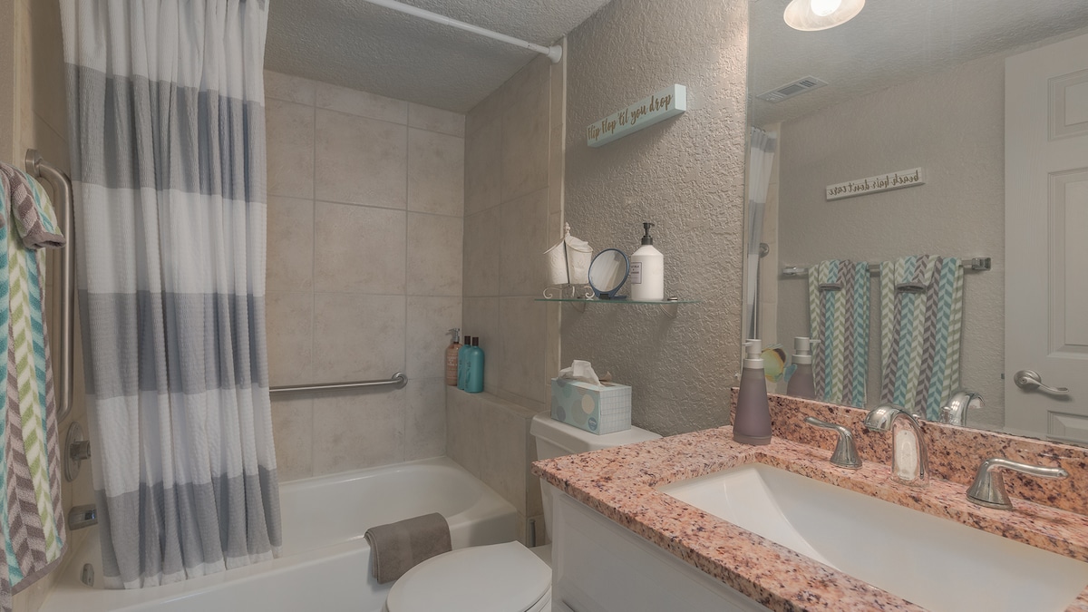 A bright bathroom features a tub and shower combination, tiled walls, and a patterned shower curtain. A granite sink counter holds neatly arranged toiletries and a decorative container. Coordinating towels hang on a rack, providing a cohesive and organized appearance.