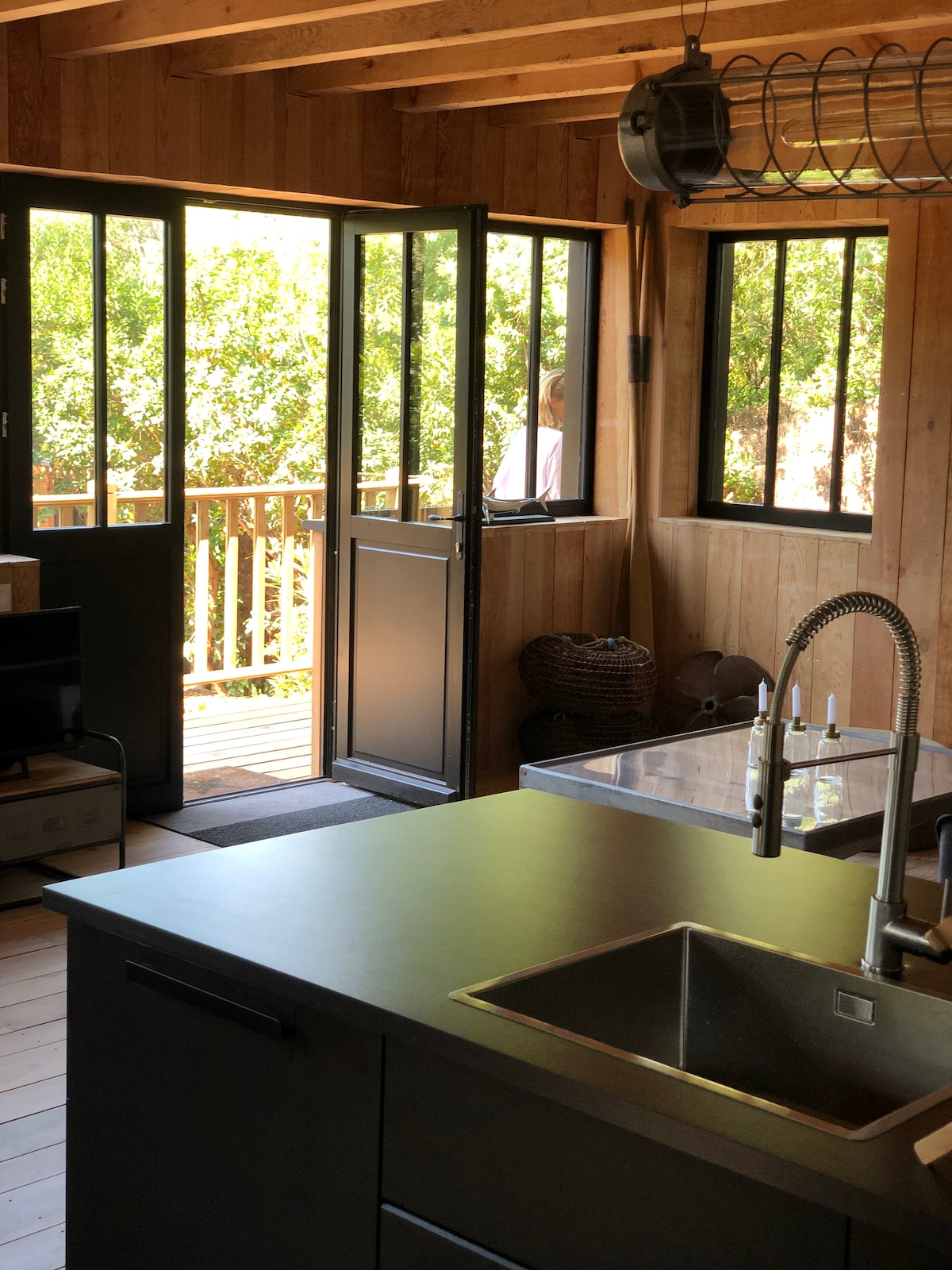 A modern kitchen is presented, featuring dark cabinetry and a stainless-steel sink. Large black-framed windows allow natural light to fill the space, with a view of a wooden terrace and greenery outside. The minimalist design highlights a functional layout with an open door leading to the outdoor area.