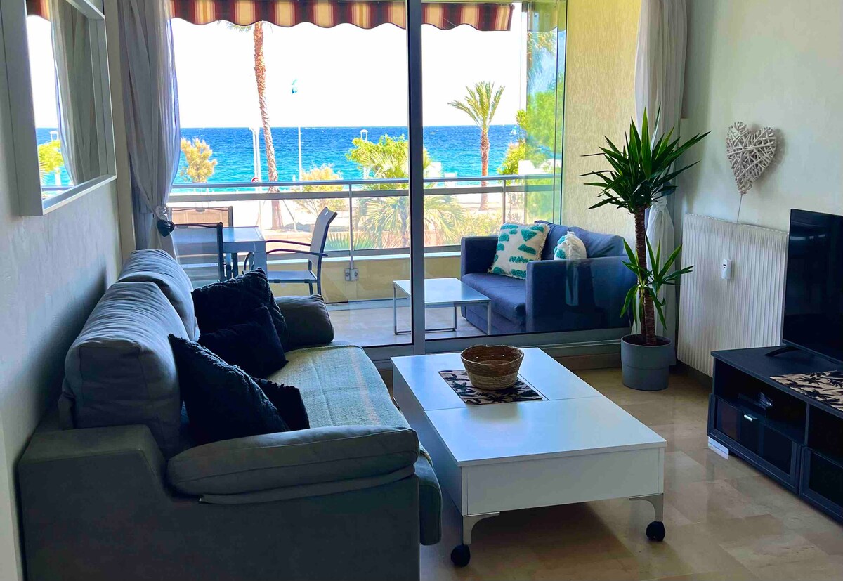 The living area features a comfortable gray sofa and armchair, facing a window with a view of the sea. A small table rests in front, and a potted plant adds a touch of greenery. Sunlight floods the space, illuminating the light-colored walls.