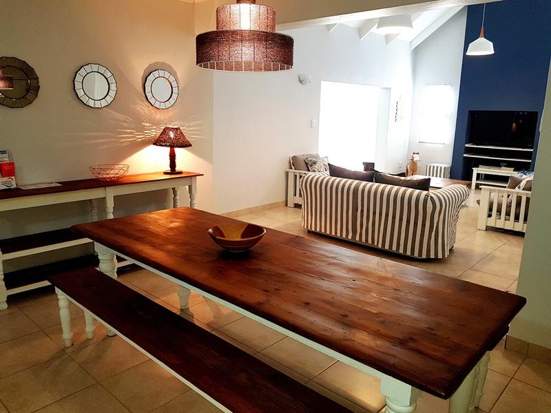 An open plan living area is shown, featuring a rustic wooden dining table with benches. A comfortable striped sofa and a TV stand are positioned in the background, well-lit by natural light coming through a large window. Decorative mirrors and a pendant light add to the inviting ambiance.