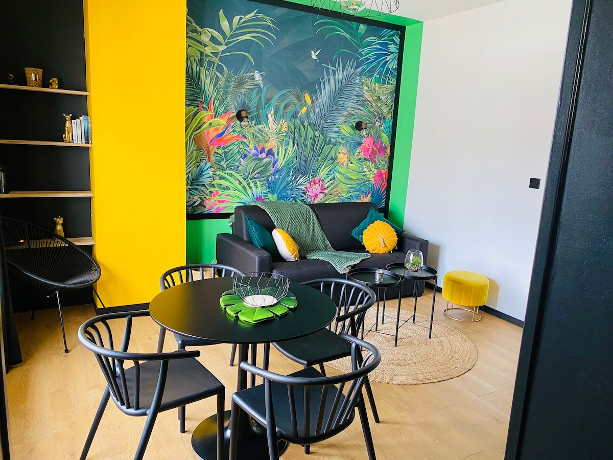 A modern living area features a black sofa adorned with green and yellow pillows. A vibrant floral mural adds color to the wall. A round black table surrounded by four chairs is situated in the center, with a woven rug and small round accent table completing the space.