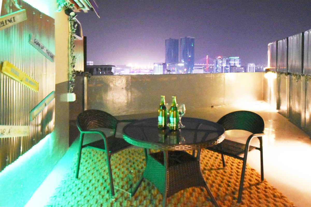 A rooftop space is featured with a table set surrounded by four chairs. Soft lighting enhances the area, providing a relaxing ambiance. A view of the city skyline is visible in the background, showcasing illuminated buildings at night.