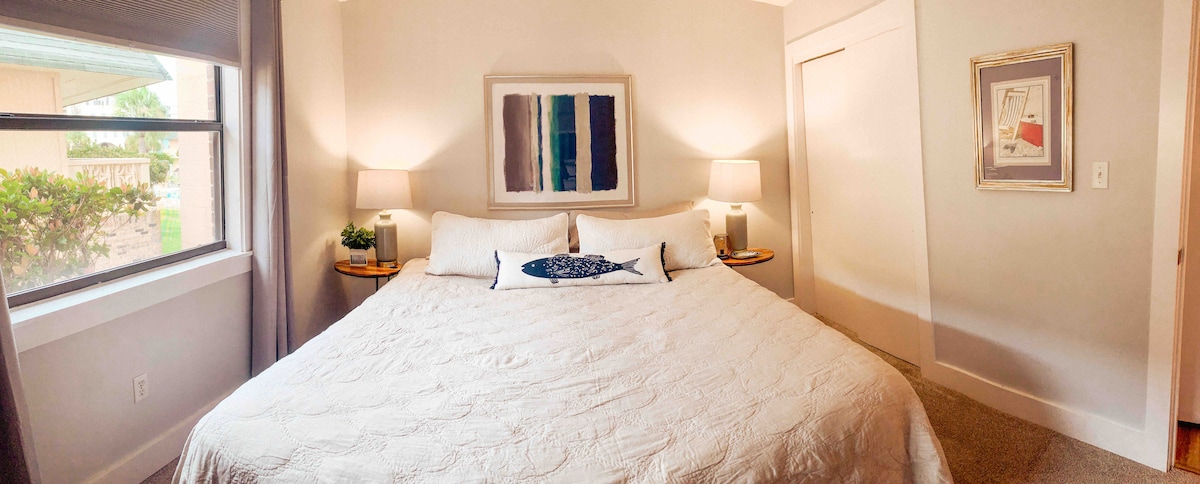 A spacious bedroom features a king bed with a light-colored quilt, flanked by two side tables with lamps. Natural light fills the room through a large window, highlighting the neutral wall tones and a piece of wall art above the bed.