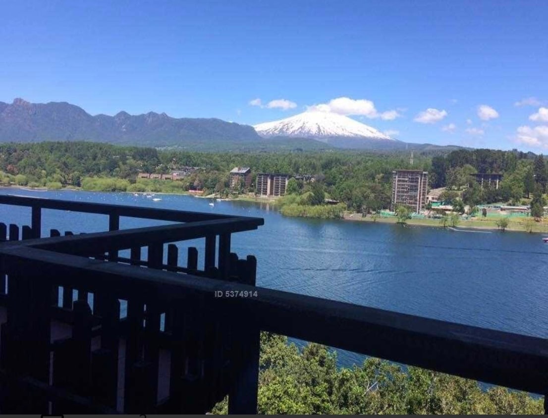 The image depicts a scenic view from a balcony overlooking a tranquil lagoon and a snow-capped volcano. Lush greenery borders the water, while a clear blue sky enhances the serene atmosphere. The perspective suggests a peaceful environment, characteristic of the natural beauty in the area.