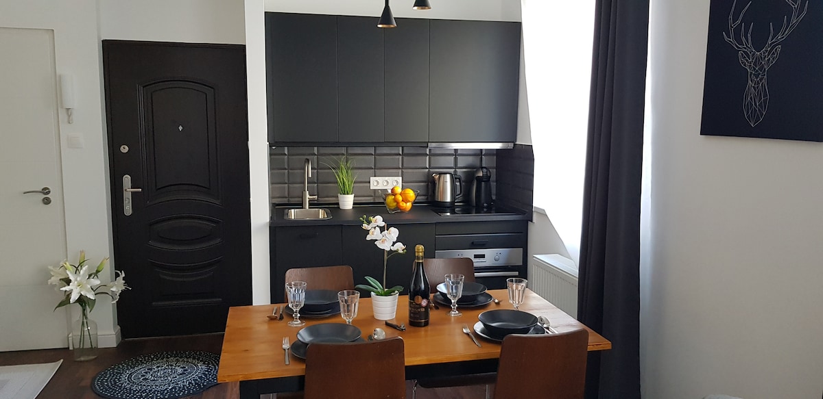 A modern kitchen area features sleek black cabinetry and a well-equipped countertop. A wooden dining table is set with tableware, complemented by a decorative flower and a bowl of fruits. Natural light filters through a partially draped window, enhancing the inviting ambiance.