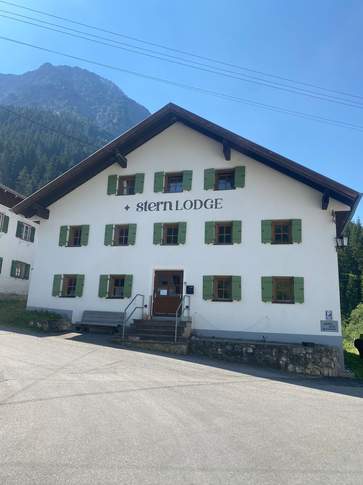 A two-story chalet is presented with a white exterior and green shutters adorning its windows. The entrance features a set of steps leading to a sturdy door, with the name 'stern LODGE' displayed prominently above. A mountainous backdrop and trees are visible in the distance.