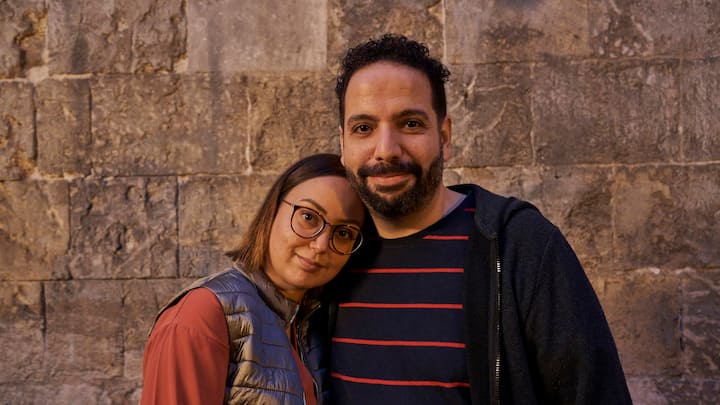 A man with a dark beard and woman with glasses stand in front of a stone wall, smiling at the camera.