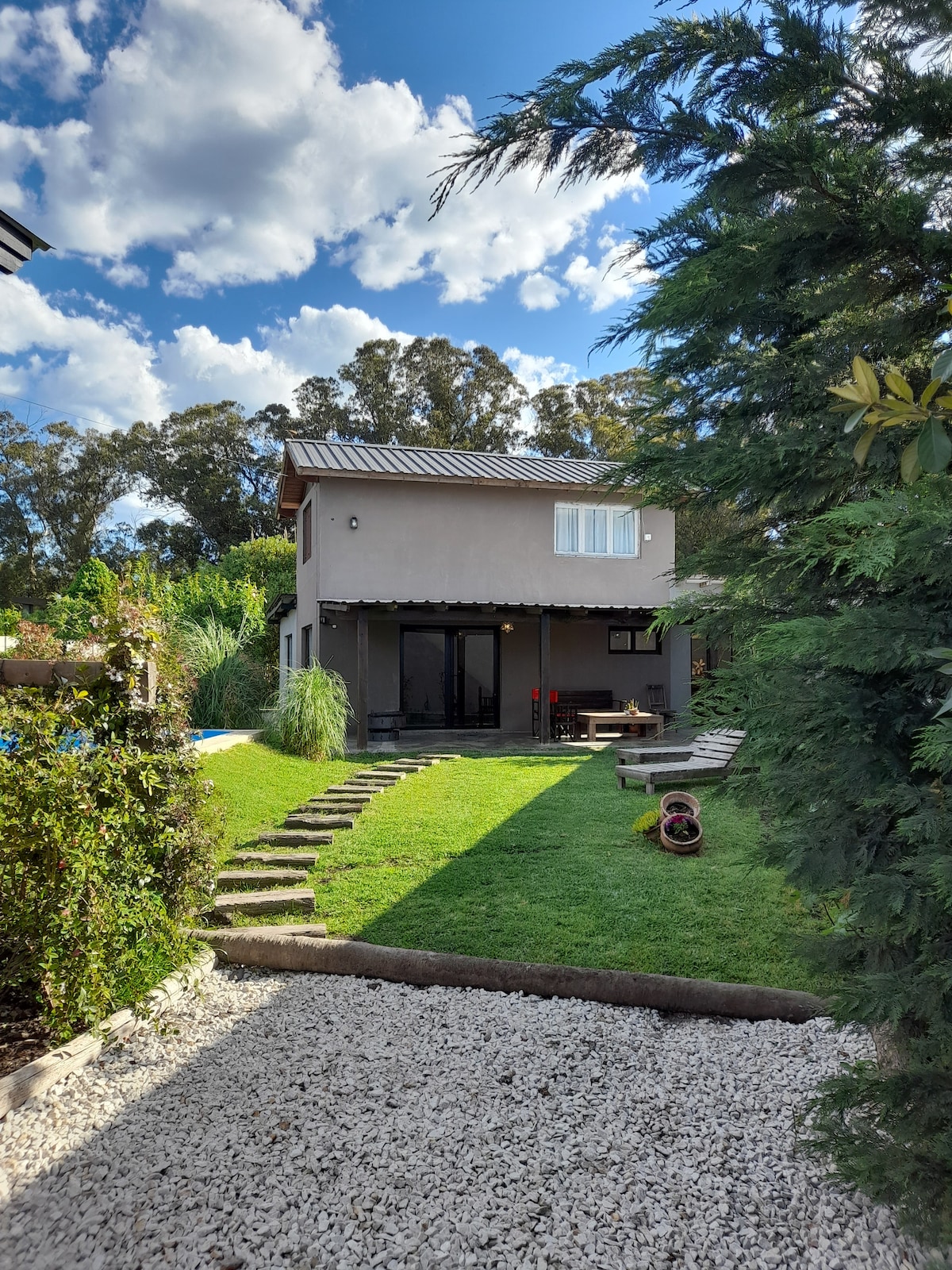 A newly constructed cabin is shown with a landscaped garden. Steps lead from a gravel path to the cabin's entrance, framed by greenery. Large windows allow natural light to enter the space, with a lawn area visible beside the cabin.