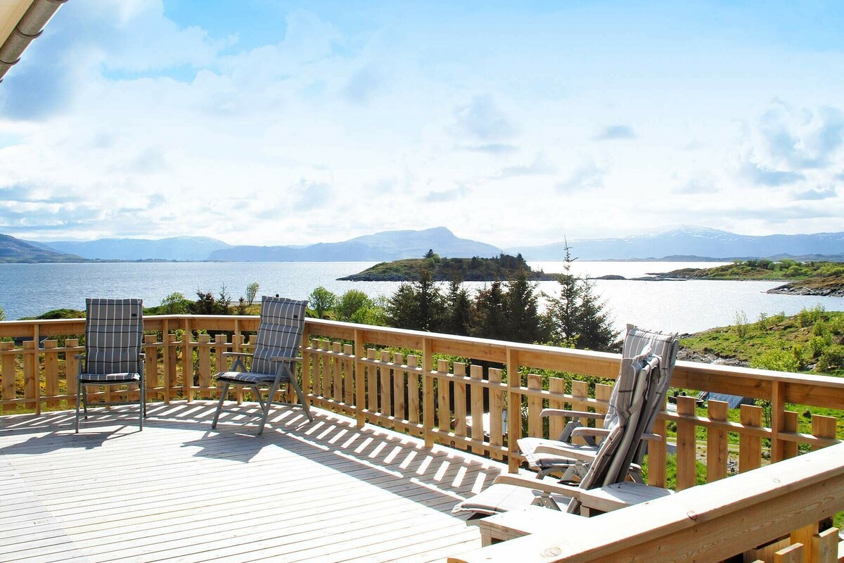 A spacious wooden deck is seen overlooking a calm water body surrounded by lush greenery and distant hills. Two comfortable chairs are positioned to take in the panoramic views, and sunlight casts soft shadows across the deck.