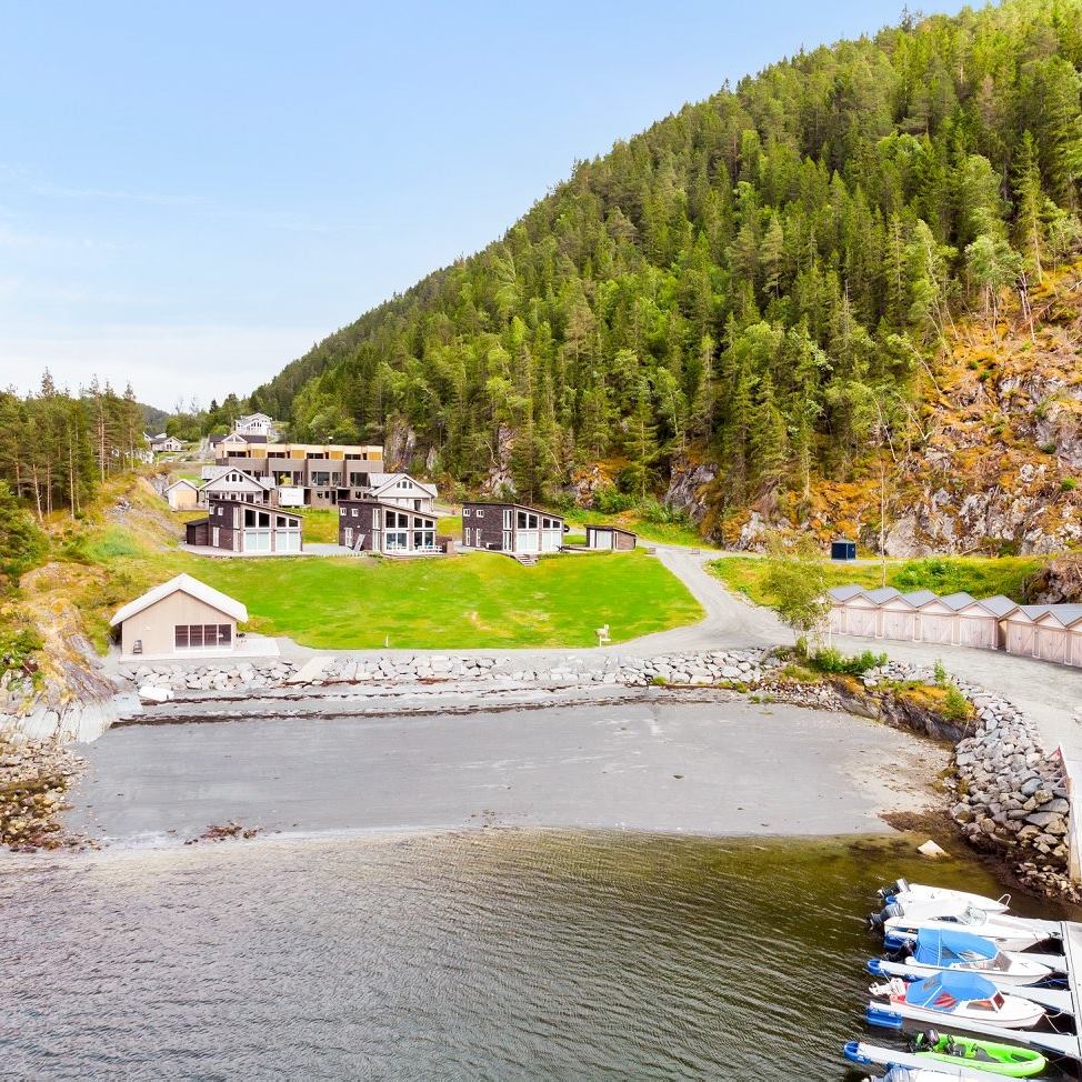 A scenic view captures the surrounding natural landscape, featuring a secluded bay with a pebbled beach. Several chalets are set back from the shoreline, framed by lush greenery and steep hills. A small marina with boats is visible along the water's edge.