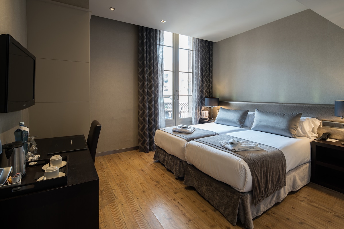 A well-appointed room features two spacious beds with neutral-toned bedding. A wooden desk and chair are positioned against the wall. Natural light floods through a large window, which is framed by curtains with a geometric pattern. A flat-screen TV is mounted on the wall.