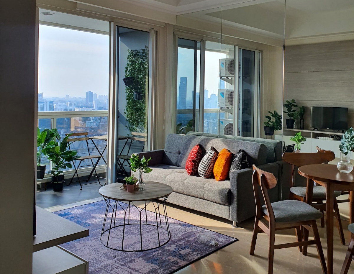 A modern living area is illuminated by natural light from large windows, showcasing a view of the city skyline. A gray sofa adorned with colorful cushions faces a round coffee table. Potted plants enhance the space, while a dining area with wooden chairs complements the contemporary design.