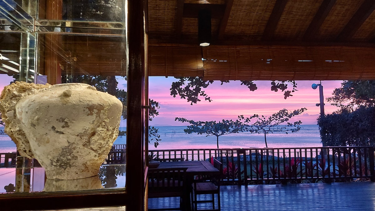 A glimpse of a serene sunset over the ocean is framed through a wooden structure. In the foreground, a rustic antique jar is displayed, while the view reveals gentle waves and silhouettes of trees against a colorful sky.
