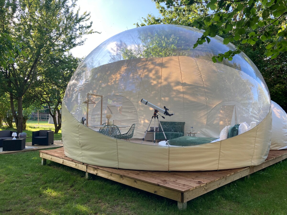 A spacious bubble structure is situated on a wooden deck surrounded by greenery. Inside, a telescope is positioned for stargazing, complemented by comfortable seating that invites relaxation. The transparent walls offer views of the natural surroundings, enhancing the outdoor experience.