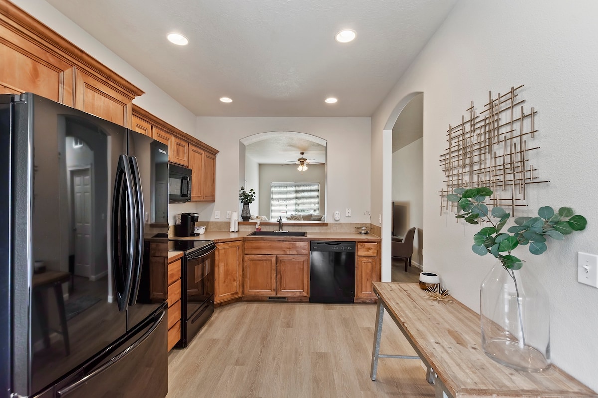 A modern kitchen features wooden cabinetry and black appliances, including a refrigerator and stove. A wooden table is positioned beside a decorative vase with greenery. Bright recessed lighting enhances the space, which connects to an adjoining area with an arched entryway.