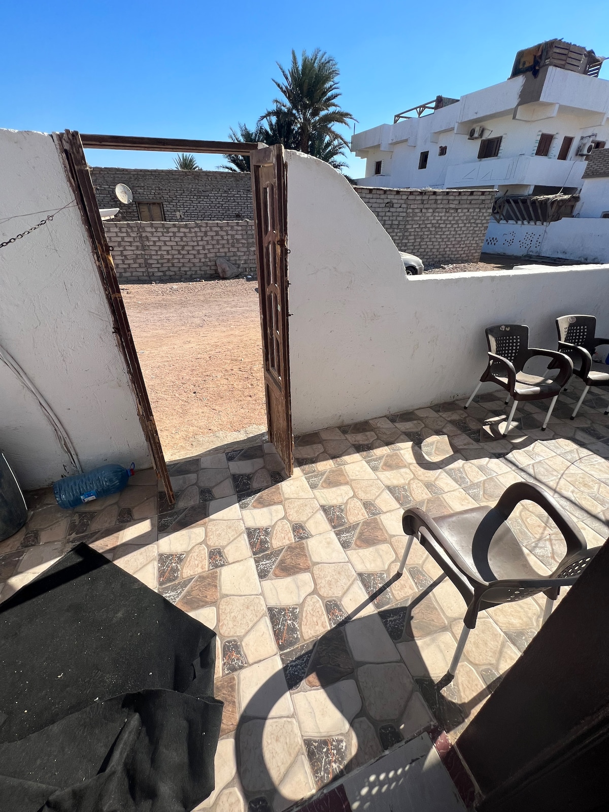 An outdoor patio is visible, featuring several black plastic chairs arranged on a patterned tiled floor. An archway leads to an open area beyond, illuminated by natural light, with hints of surrounding structures and vegetation in the background.