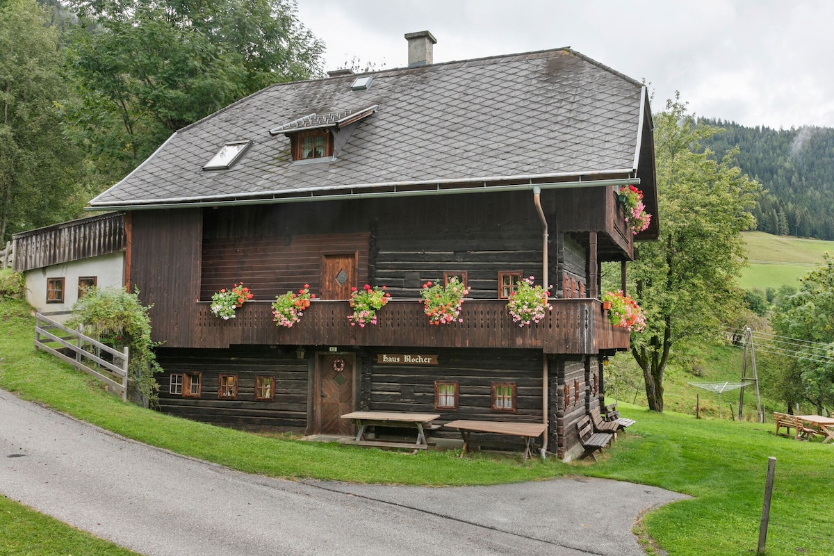 The holiday home features a charming wooden exterior with numerous flower boxes adorning the windows. The building is surrounded by lush greenery and a well-maintained lawn, with a gravel path leading up to the entrance. A covered balcony offers additional outdoor space.