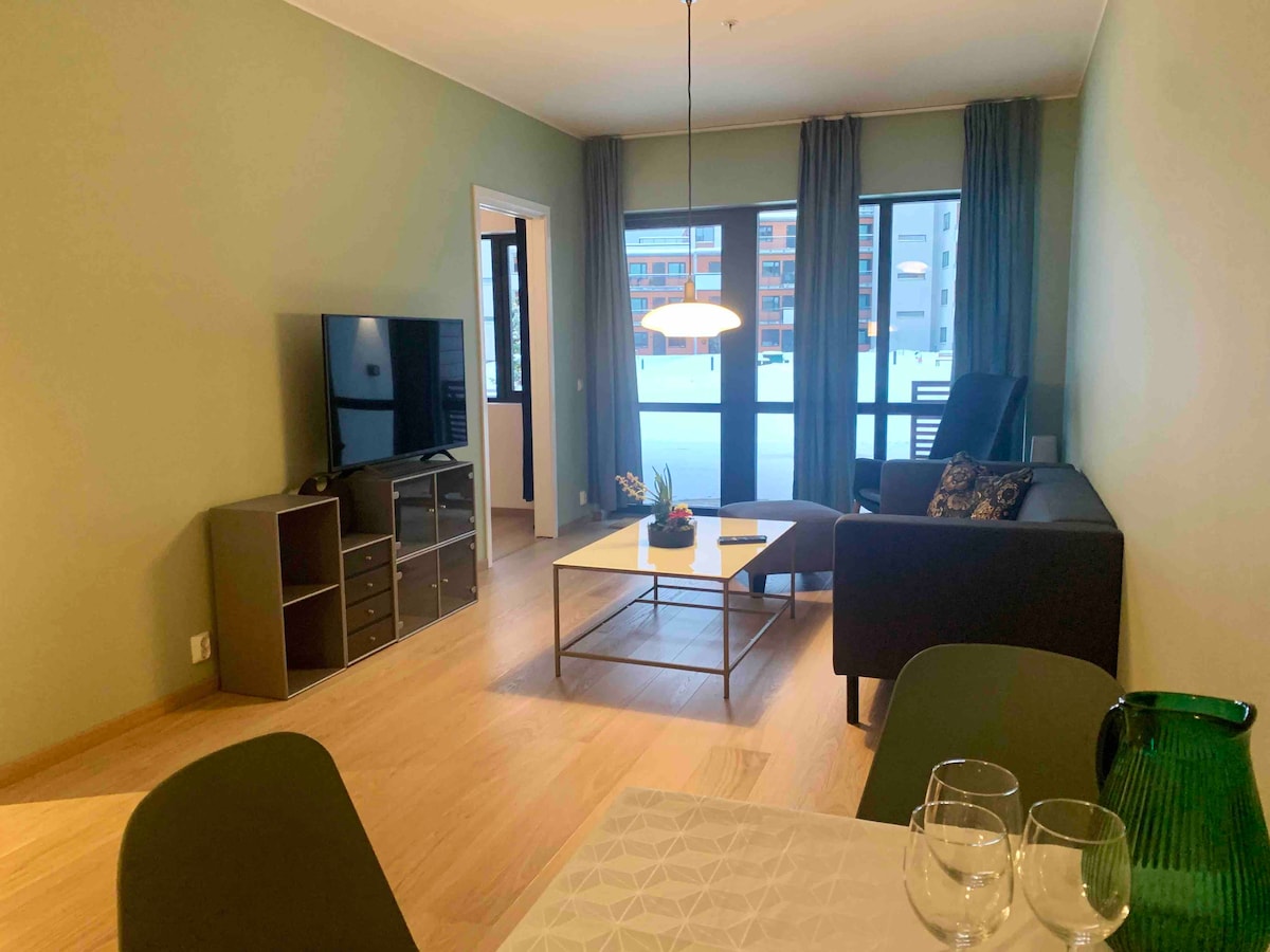 A comfortable living area showcases a modern seating arrangement with a dark sofa and a coffee table. A television is set on a stand, while large windows allow natural light to brighten the space. Elegant curtains frame the windows, enhancing the room's warmth.