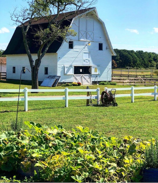 A large white barn with a distinctive peaked roof is framed against a clear blue sky. Lush green grass surrounds the building, while a section of garden with various plants is visible in the foreground. A vintage plow rests nearby on the well-maintained property.