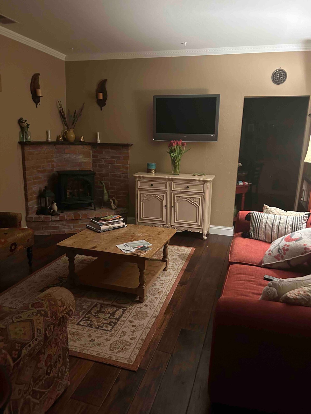 A cozy living room is showcased, featuring a red sofa facing a rustic brick fireplace. A flat-screen TV is mounted on the wall above the fireplace. A wooden coffee table sits in the center, adorned with magazines. Warm lighting creates a relaxing ambiance.