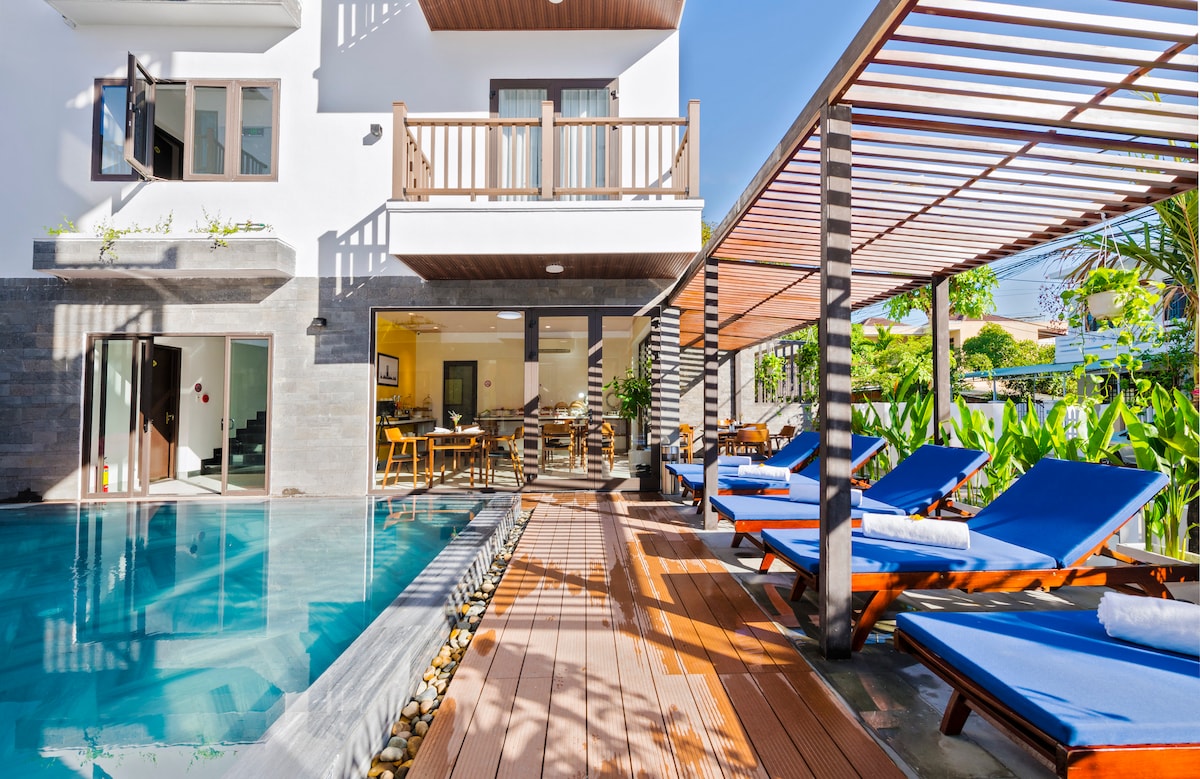 An outdoor area showcases a swimming pool with clear water bordered by wooden decking. Sun loungers with blue cushions are positioned beside the pool, while a shaded seating area is seen in the background. Lush greenery and plants surround the space, enhancing the serene setting.