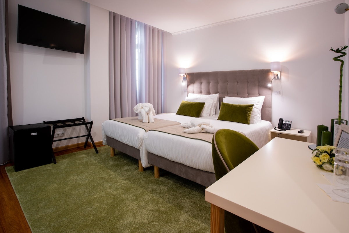 A well-appointed guest room is presented with two spacious beds featuring subtle green accents. Soft lighting is provided by wall sconces, and a modern television is mounted on the wall. A small desk and a luggage rack are included, along with a green area rug layering the floor.