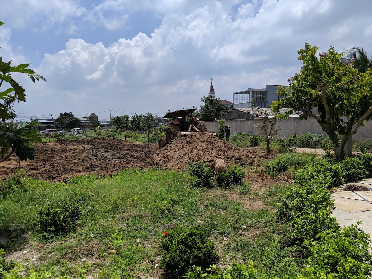 A spacious outdoor area is shown, featuring freshly tilled soil surrounded by patches of grass and scattered green bushes. The sky is bright with scattered clouds, and a structure in the distance appears to be a church steeple among low buildings.