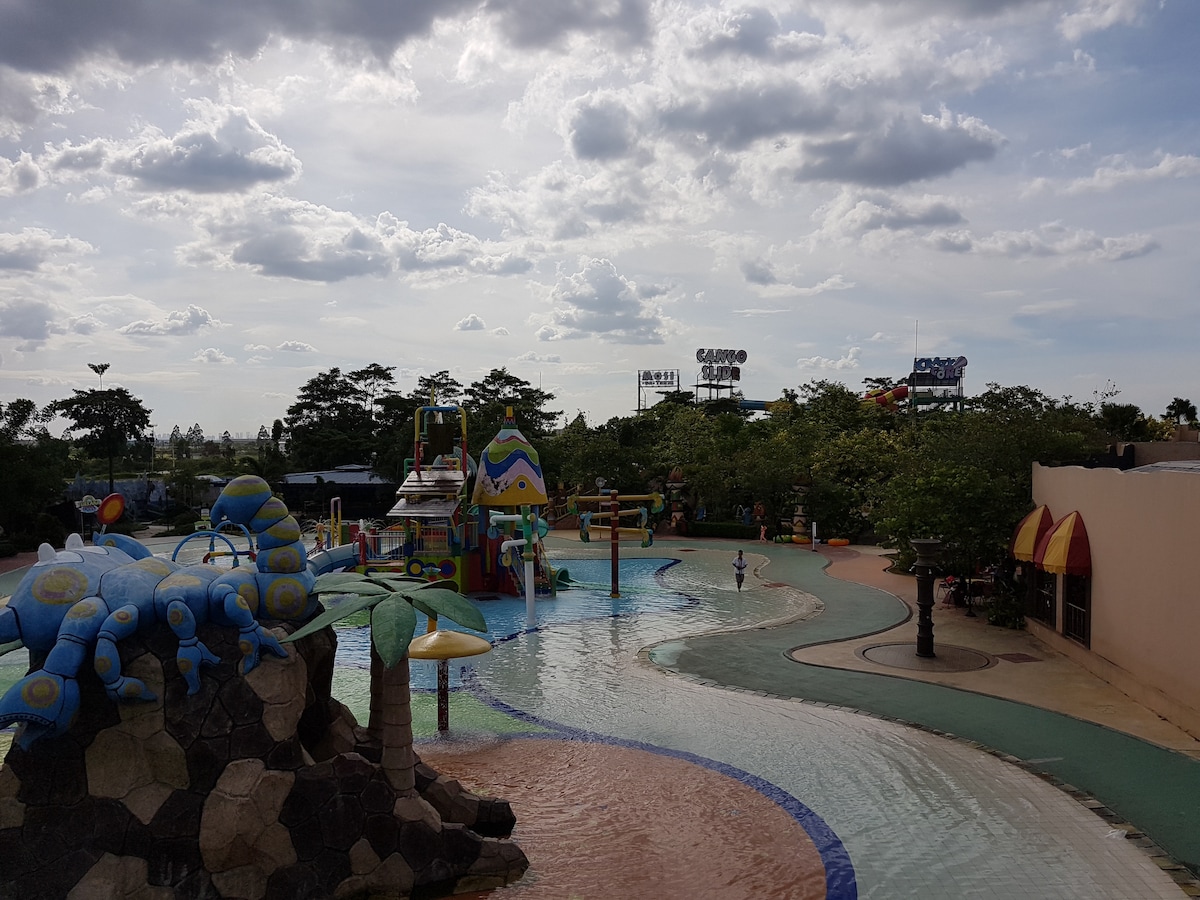 A colorful waterpark is showcased, featuring various water attractions and playful structures. The landscape includes gentle slides and playful sculptures, surrounded by lush greenery. The atmosphere appears lively, with scattered poolside seating and the distant outlines of buildings under a partly cloudy sky.