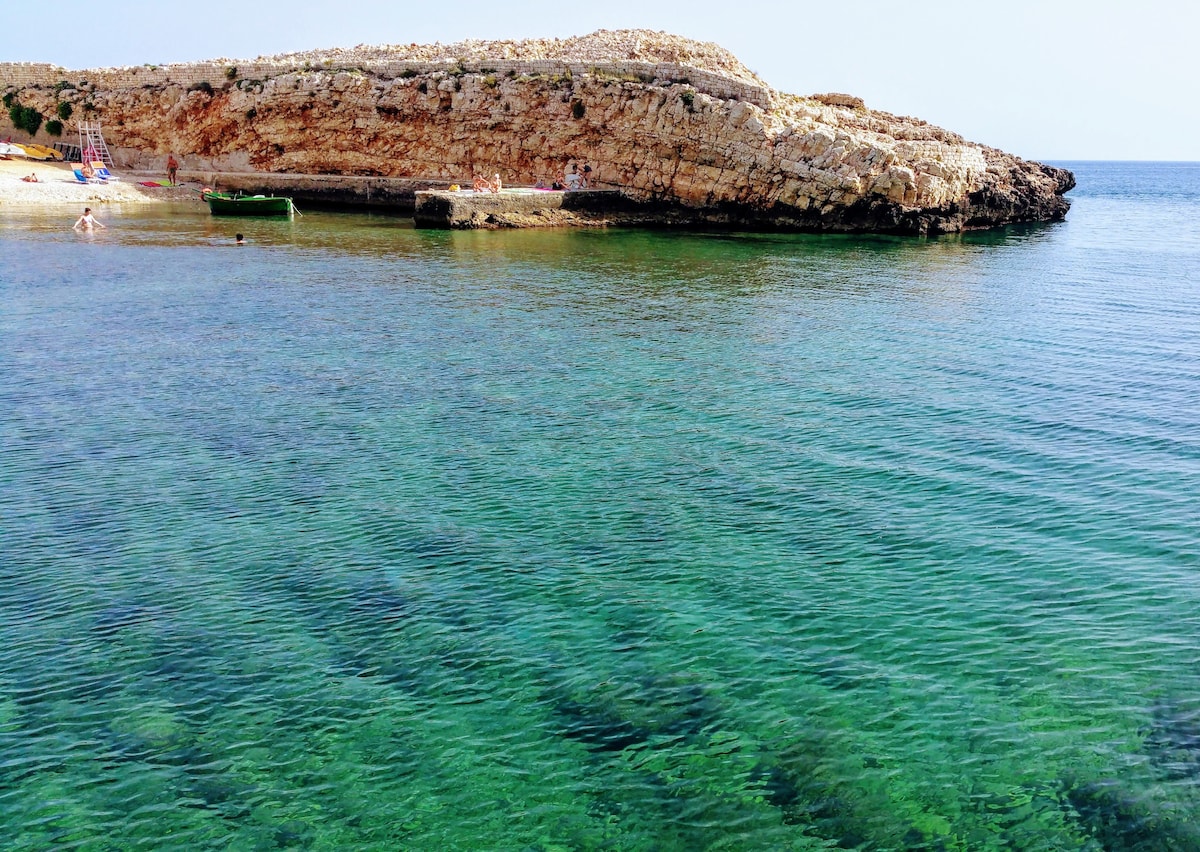 A clear view of a tranquil coastal scene featuring calm, turquoise waters gently lapping against a rocky shoreline. The rocky outcrop is visible in the background, with people enjoying the beach and swimming in the distance.
