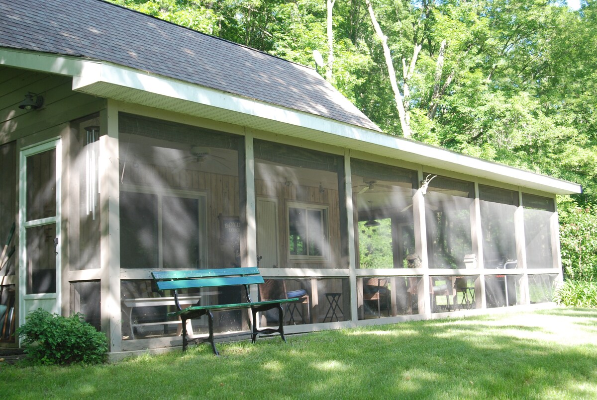 A large screened porch offers shade and views of the surrounding greenery. It features multiple seating areas, including a bench, providing a comfortable space for relaxation. Sunlight filters through the trees, creating a serene atmosphere in the outdoor area.