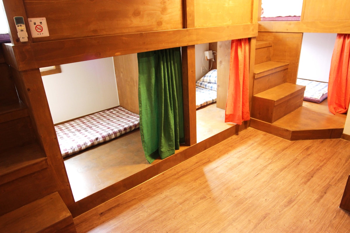 A shared dormitory space is shown, featuring wooden framing and steps leading to elevated sleeping areas. Each bed is separated by colorful curtains, offering privacy while ample natural light illuminates the room. The floor is finished with warm, wooden planks, enhancing the cozy ambiance.