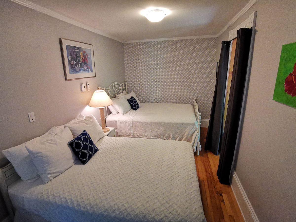 A well-lit bedroom features two beds, one queen size and one double size, neatly made with white linens. A bedside lamp provides soft illumination. Artwork adorns the walls, adding a touch of color, while curtains frame a window, allowing natural light to enter.