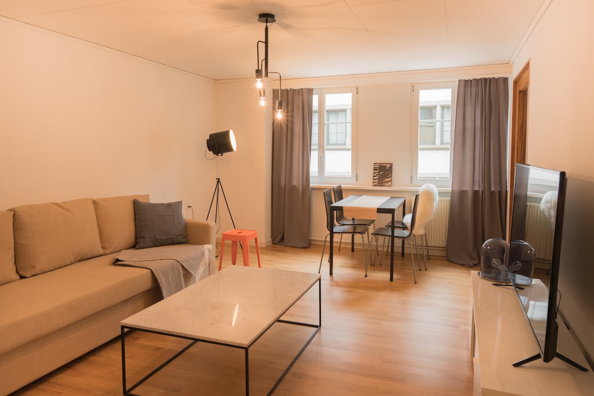 A bright living area is displayed, featuring a soft sofa positioned next to a sleek marble coffee table. A dining table with chairs is situated nearby, illuminated by natural light filtering through large windows, which are framed by flowing gray curtains.
