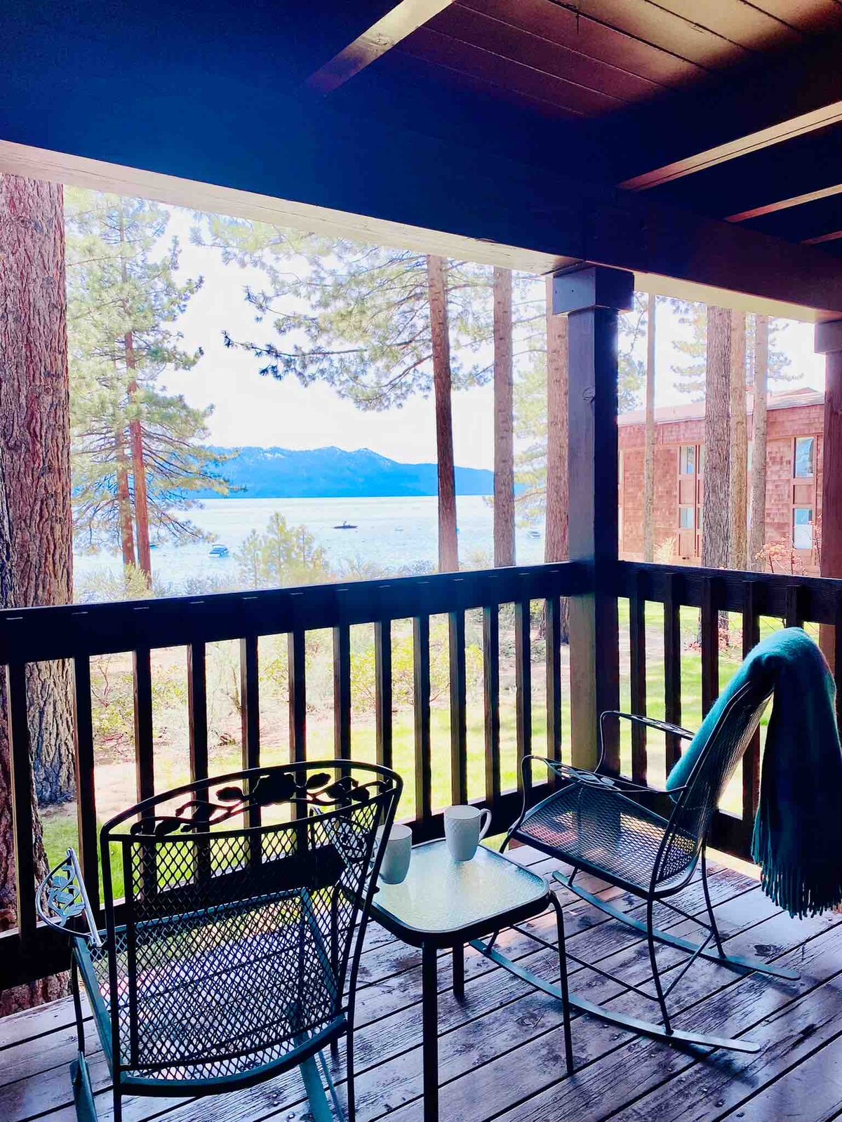 A balcony offers views of the alpine lake and pine trees. Two metal chairs and a small table are visible, with a blanket draped over one of the chairs. The scene is framed by the balcony's wooden railing, creating a serene outdoor space.