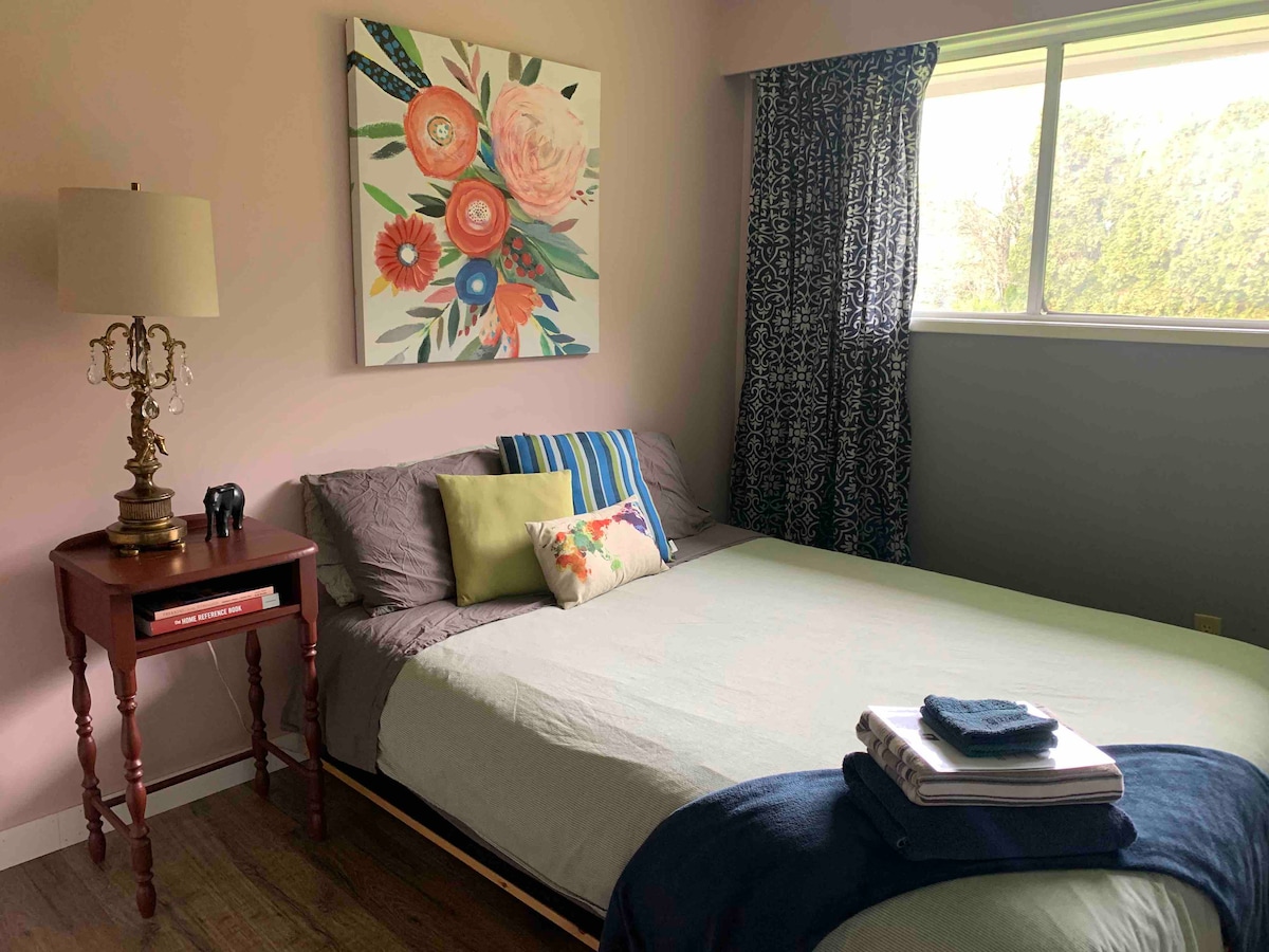 A guest room features a spacious bed with various decorative pillows, set against a soft pink wall. A large floral artwork adorns the wall above the bed. A bedside table with a lamp and a small decorative figure is positioned beside the bed. Natural light enters through the window, partially covered by patterned curtains.