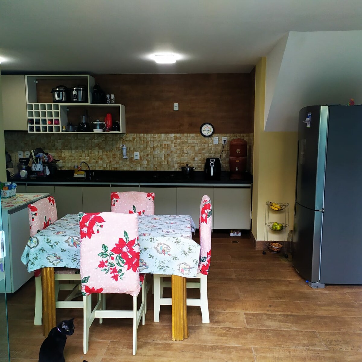 A well-appointed kitchen features a dining table surrounded by chairs with floral tablecloths. Modern appliances, including a coffee maker and a refrigerator, are prominently displayed. A clock is mounted on the wall, and a small cat is seated on the floor, adding a homey touch.