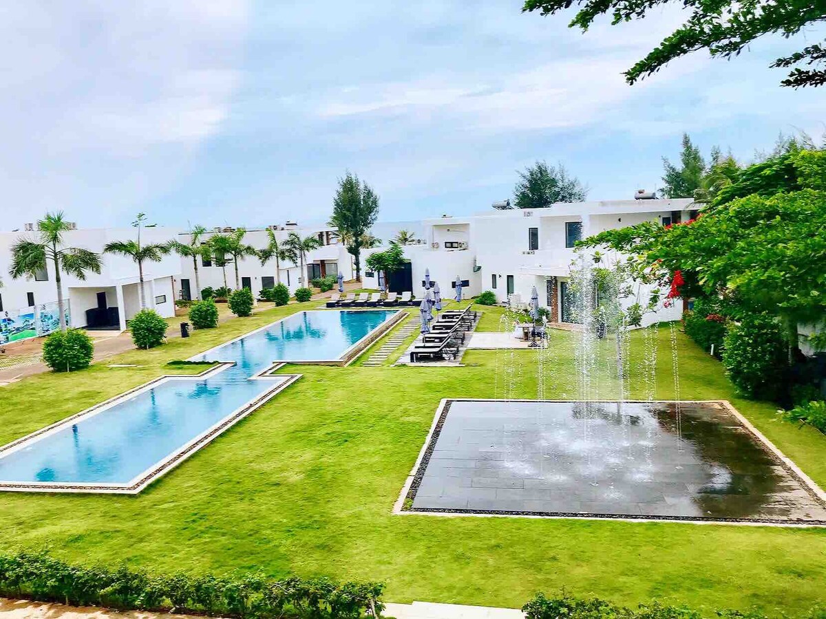 A landscaped area is shown with two swimming pools surrounded by lounge chairs. Neatly trimmed grass and ornamental trees add to the green space. A water feature provides a gentle flow of water, enhancing the outdoor environment. White structures are visible in the background.